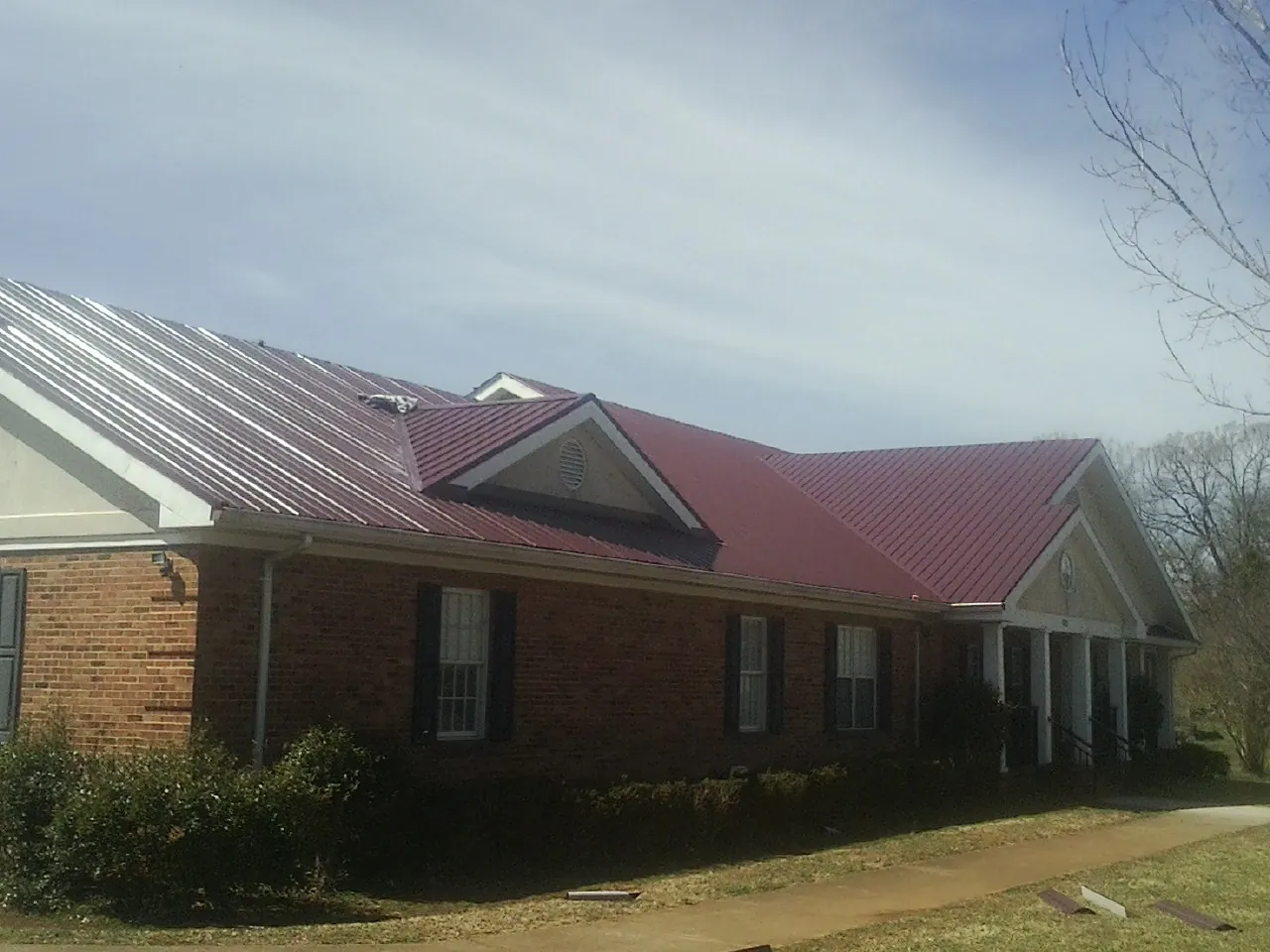 Red metal roof with gabled entries on a long brick building, from a Hail Damage Roof Repair job in Syria