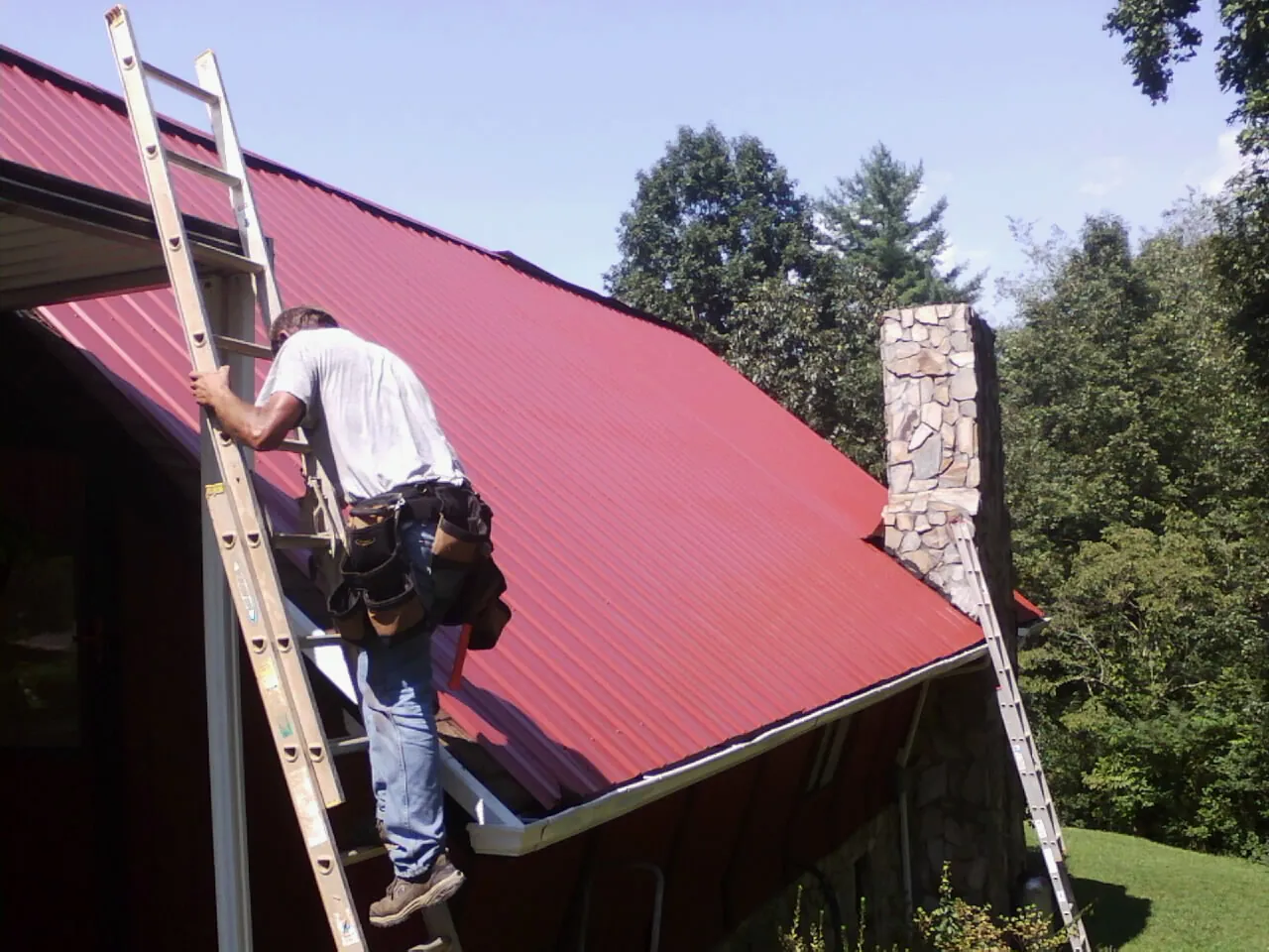 Roofer on a ladder beside a red metal roof and stone chimney — Soffit & Fascia Repair by Roofing Brothers in Syria
